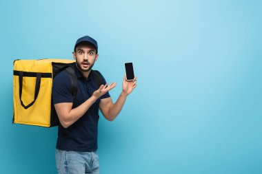 Şok olmuş Müslüman kurye termo çantayla akıllı telefonu işaret ediyor ve mavi ekranda boş ekranı var.