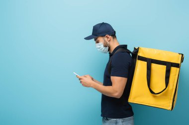 Tıbbi maskeli Arap kuryenin yan görüntüsü mavi cep telefonundaki sırayı kabul ediyor