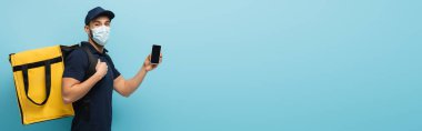 Tıbbi maskeli Arap kurye, mavi pankartta boş ekranlı akıllı telefon gösteriyor.