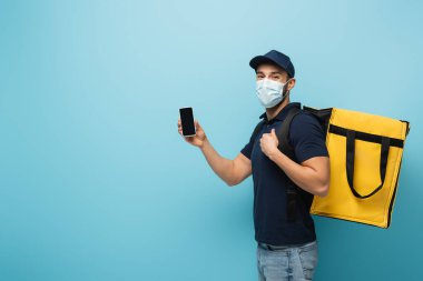 Maskeli Müslüman kurye mavi ekranda boş ekranlı cep telefonunu gösteriyor.