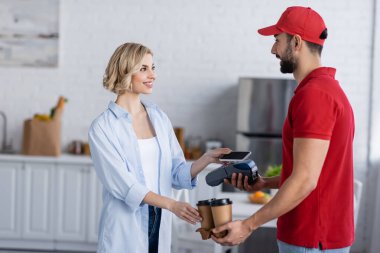 Müslüman kurye ödeme terminalini ve kağıt bardakları cep telefonlu neşeli kadının yanında tutuyor.