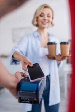 Teslimatçı adam ödeme terminalini bulanık arka planda akıllı telefonlu gülümseyen kadının yanında tutuyor.