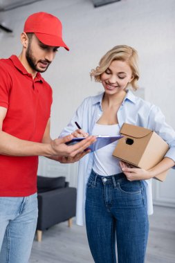 happy blonde woman holding parcel and writing on clipboard near muslim postman clipart