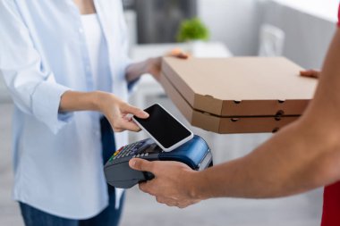 Ödeme terminalini elinde tutan teslimatçı ve cep telefonu olan bir kadının yanında pizza kutuları.