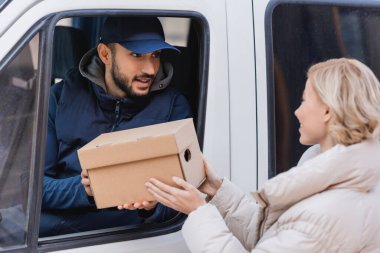 muslim postman in truck giving parcel to blonde woman on blurred foreground clipart
