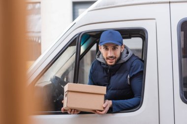 positive delivery man holding parcel while looking out car window on blurred foreground clipart