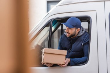 muslim delivery man with carton box looking out car window on blurred foreground clipart