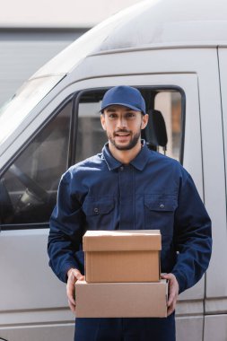 Neşeli müslüman postacı dışarıda arabanın yanında dururken elinde paketler tutuyor.