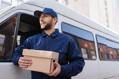 happy arabian postman looking away while standing with parcel near car clipart