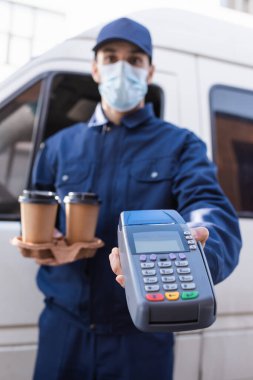 Tıbbi maskeli Arap kurye arka planda bulanık bir ödeme terminaline gitmek için elinde kahve tutuyor.