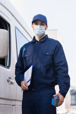 arabian postman in protective mask standing near car with payment terminal and clipboard clipart