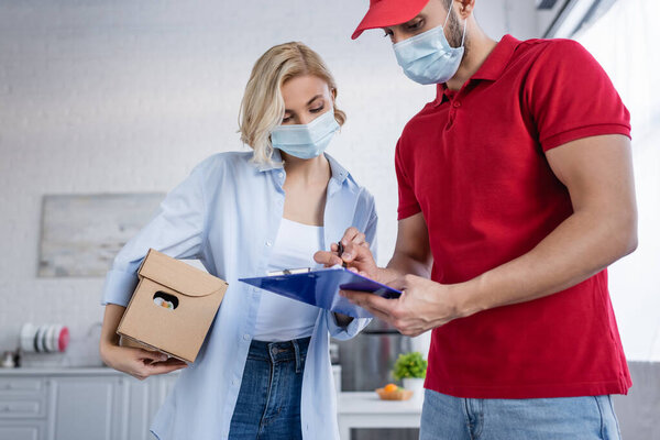 muslim delivery man writing on clipboard near woman in medical mask holding parcel 