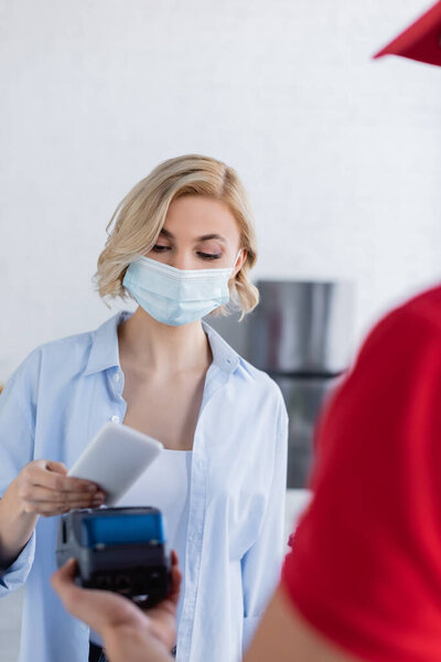 delivery man on blurred foreground holding payment terminal near woman in medical mask, blurred foreground