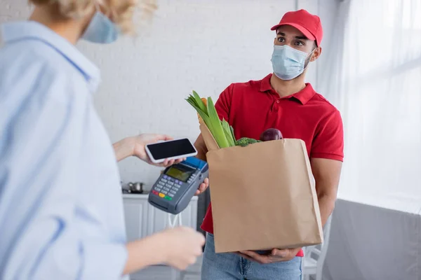 arabian man holding payment terminal and paper bag with food near woman with smartphone on blurred foreground