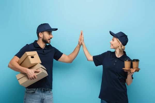 веселые мультикультурные курьеры с бумажными стаканчиками и коробками, дающие пять на голубой