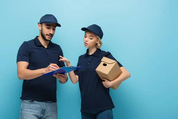 muslim delivery man writing order near colleague pointing with hand isolated on blue