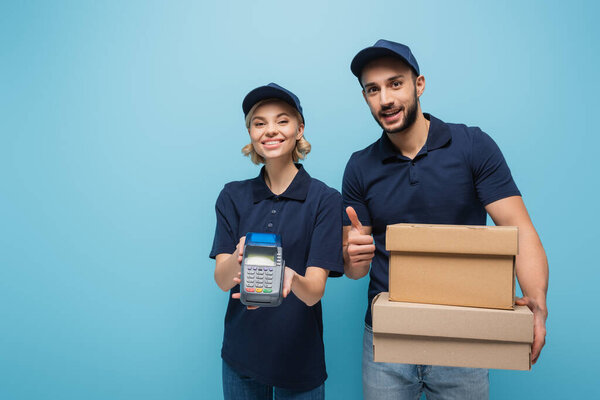 happy arabian delivery man showing thumb up near colleague with payment terminal on blue
