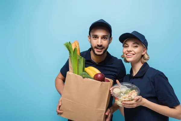 cheerful multicultural couriers with food in packages smiling at camera isolated on blue
