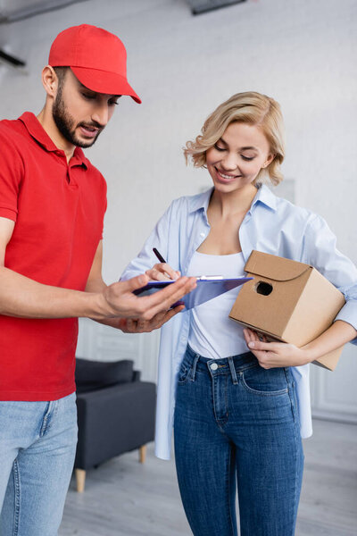 happy blonde woman holding parcel and writing on clipboard near muslim postman