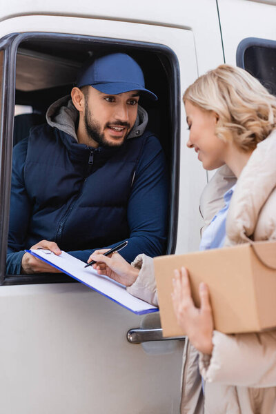 blonde woman signing order on clipboard while holding parcel near smiling arabian postman, blurred foreground
