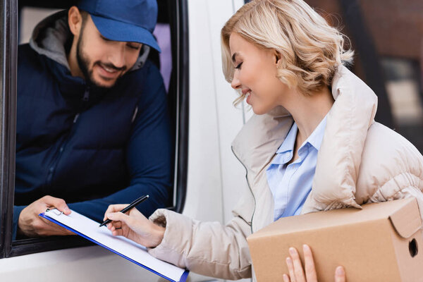 joyful woman signing order while holding parcel near arabian postman in truck on blurred background