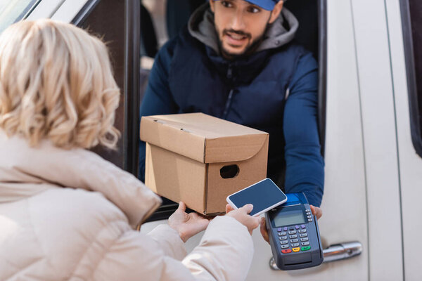 arabian delivery man giving payment terminal and parcel to woman with cellphone on blurred foreground