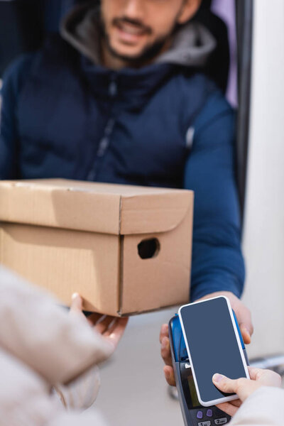 cropped view of woman holding parcel and paying with smartphone on terminal near postman on blurred background