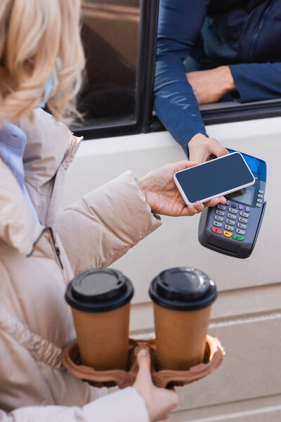 cropped view of woman holding paper cups and smartphone near payment terminal on blurred foreground