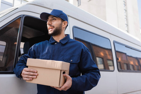 happy arabian postman looking away while standing with parcel near car