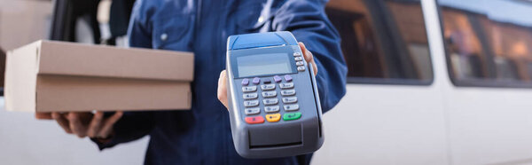 cropped view of delivery man with carton box and payment terminal on blurred background, banner