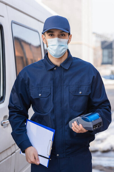 arabian courier in medical mask holding payment terminal and clipboard while standing near car