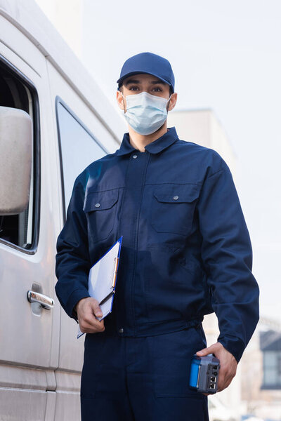 arabian postman in protective mask standing near car with payment terminal and clipboard