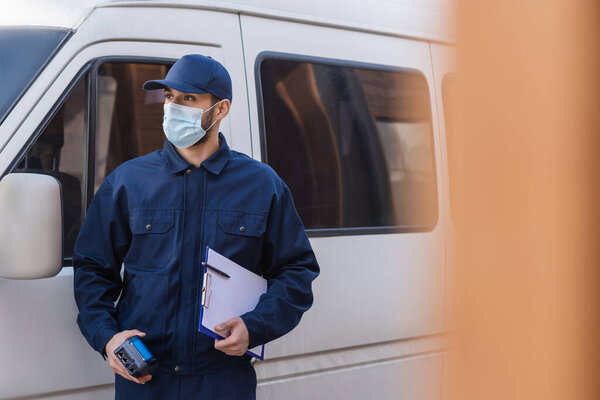 muslim delivery man in medical mask looking away while standing near car on blurred foreground