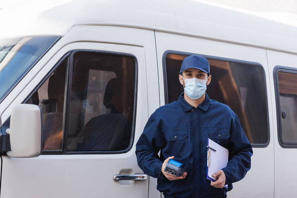 young arabian courier in medical mask standing with clipboard and credit card reader near car