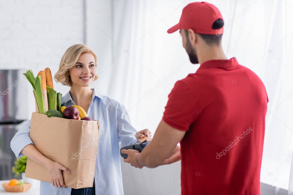 arabian delivery man holding terminal near blonde woman with fresh food in paper bag