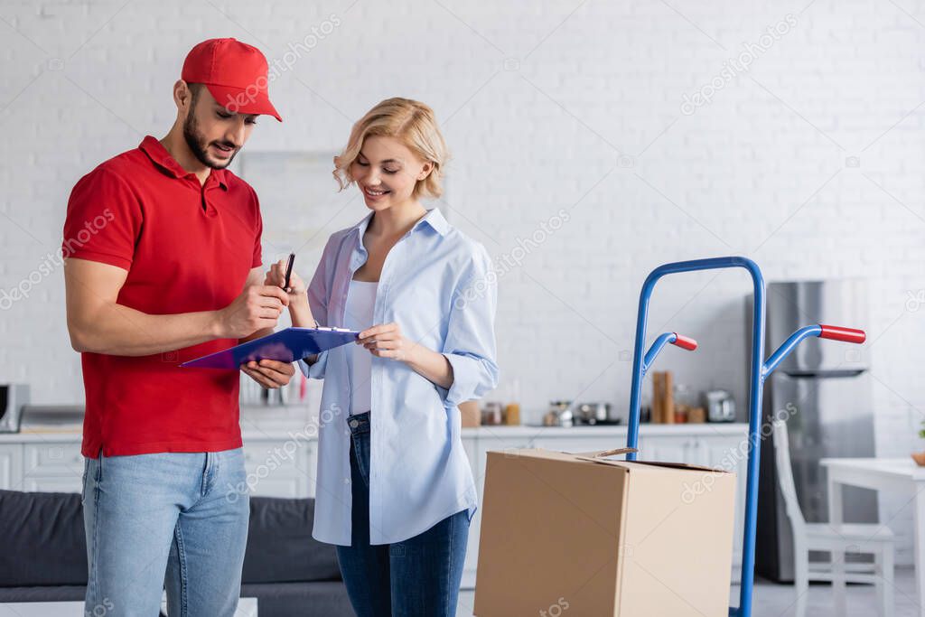 muslim postman giving pen and clipboard to joyful woman near cart with parcel