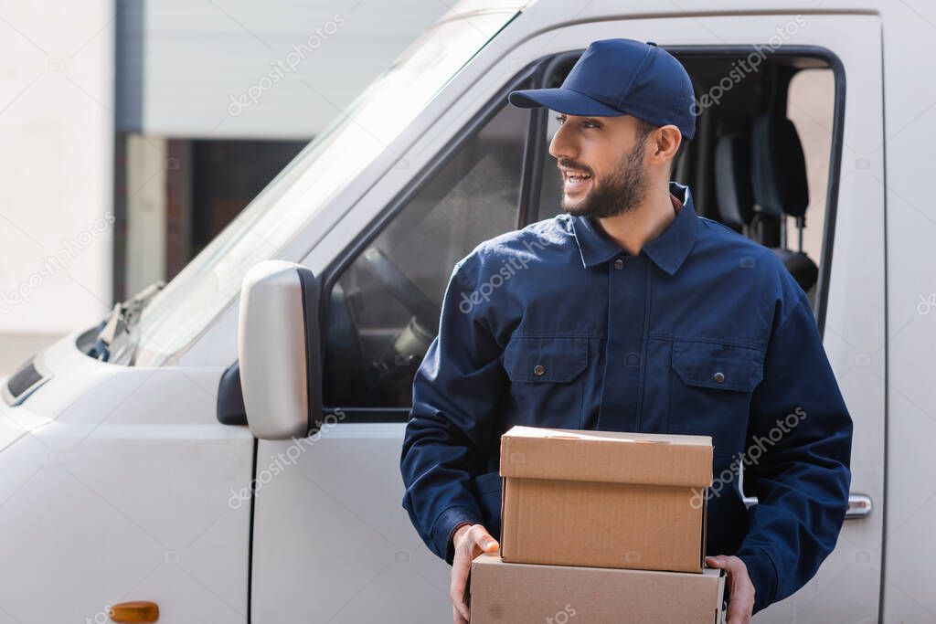 smiling arabian courier looking away while holding parcels near car