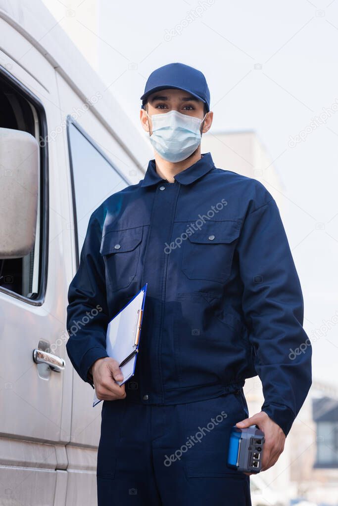 arabian postman in protective mask standing near car with payment terminal and clipboard