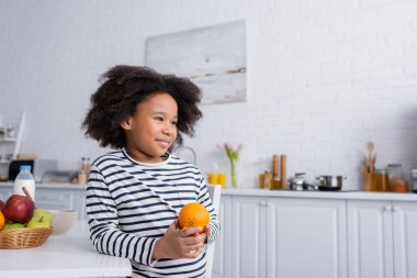 Gülümseyen Afrikalı Amerikalı çocuk taze portakalla mutfak masasının yanında duruyor.
