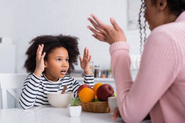 Heyecanlı Afrikalı Amerikalı çocuk kahvaltıda bulanık ön planda annesiyle konuşurken el kol hareketi yapıyor.