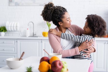Neşeli Afro-Amerikan anne ve kızı mutfakta taze meyvelerin yanında bulanık ön planda kucaklaşıyorlar.