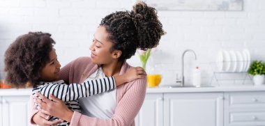 Mutlu Afro-Amerikan anne ve kızı mutfakta sarılırken birbirlerine bakıyorlar.