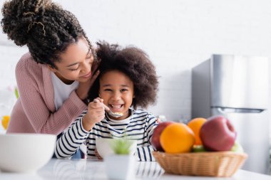 Afro-Amerikan kadın mutlu çocuğun saçlarına dokunuyor meyvelerin yanında mısır gevreği yiyor bulanık ön planda