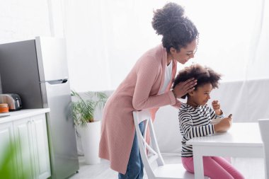 Genç Afrikalı Amerikalı kadın, mutfakta kahvaltı yapan çocuğun saçına dokunuyor.