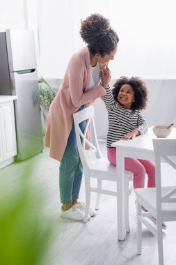 Neşeli Afrikalı Amerikalı kız mutfakta kahvaltı ederken annesinin yüzüne dokunuyor.
