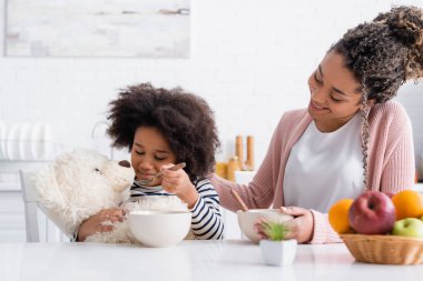 african american woman smiling near daughter feeding teddy bear with corn flakes clipart