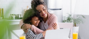 happy african american mom and daughter embracing while watching film on laptop, blurred foreground, banner clipart