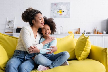 Mutlu Afrikalı Amerikalı çocuk ve anne evde kanepede otururken kucaklaşıyorlar.