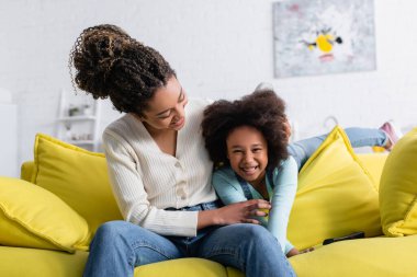 happy african american woman tickling excited daughter on sofa at home clipart