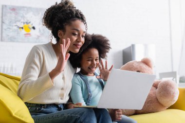 Gülümseyen Afro-Amerikan anne ve çocuğu laptopta görüntülü sohbet sırasında el sallıyorlar.